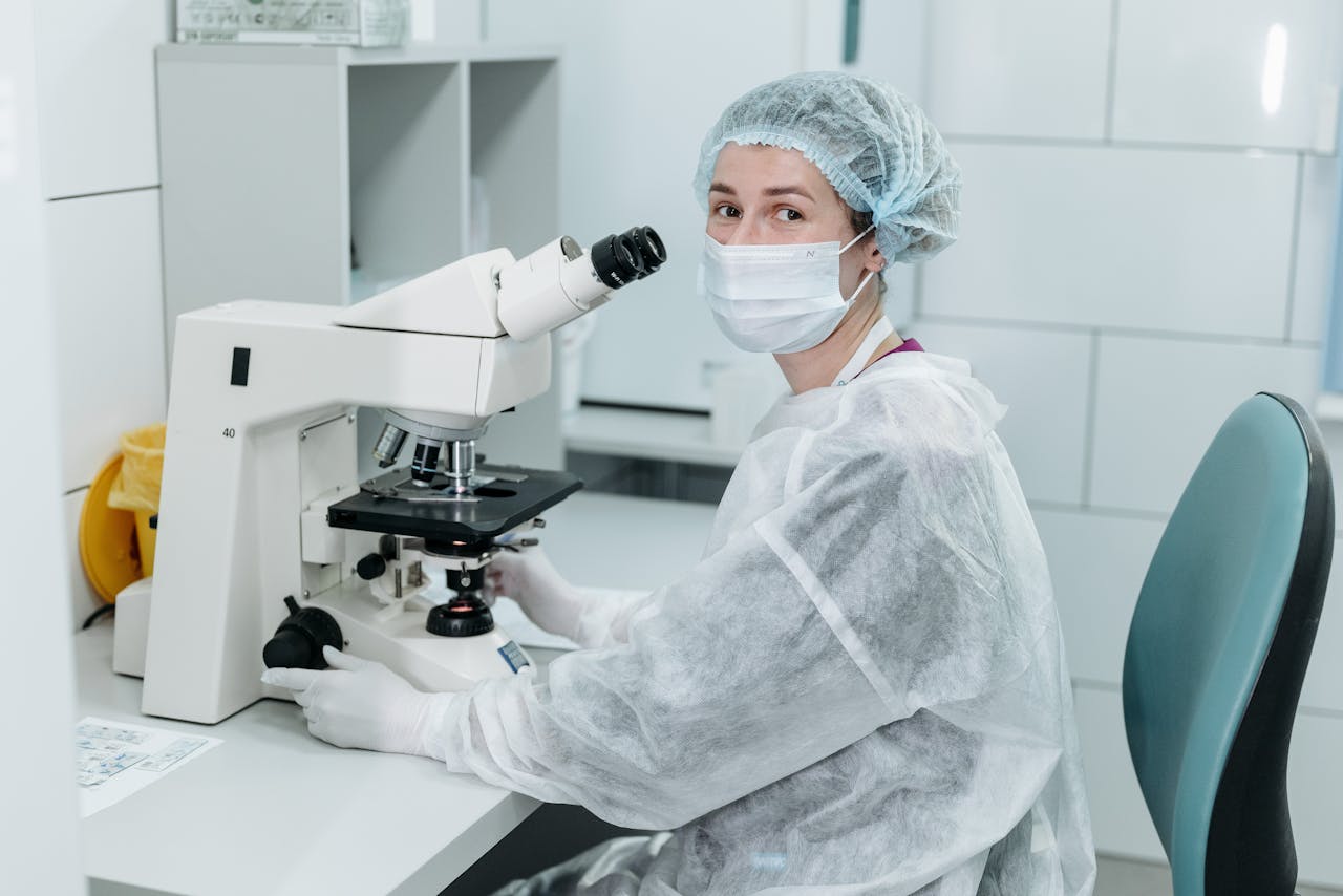 Scientist working with microscope in sterile lab environment, wearing protective gear and a mask.